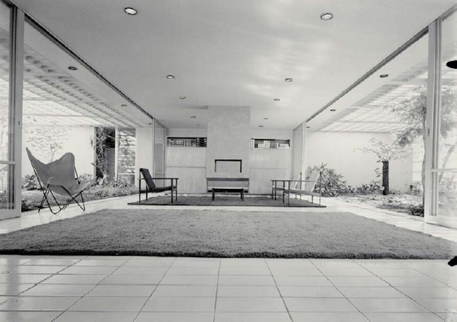 a imagem ilustra uma foto de um ambiente interno de uma residência. Há uma sala ampla, de piso frio, dois tapetes, uma cadeira próxima ao primeiro tapete. Sobre o segundo tapete há uma mesa de centro, duas poltronas à direita e uma à esquerda. Ao fundo, dividindo o ambiente, há uma estrutura que vai do teto ao chão com dois armários com livros.