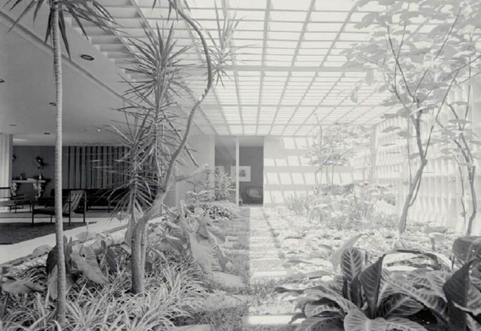 a imagem ilustra uma foto de um jardim interno de uma residência localizado ao lado da sala de estar. O jardim é amplo e iluminado, com arbustos e folhagens em suas laterais. Ao centro há um piso rústico. Seu teto é de pergolado. Sua integração com a sala de estar é através de uma porta ampla de vidro, na lateral. 